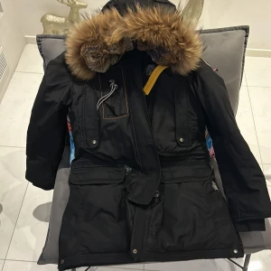 Svart parkajacka med pälsdetaljer från Parajumpers - Snygg svart parkajacka från Parajumpers med pälsfodrad huva och flera praktiska fickor. Jackan har en dragkedja och knappar framtill samt en gul remdetalj vid kragen. Perfekt för kalla vinterdagar. Inne i luvan är den skadad som ni kan se på en av bilderna. Priset går att diskuteras