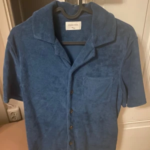 Blå frottéskjorta från Studio Total - Säljer en blå frottéskjorta från Studio Total med korta ärmar och knappar framtill. Skjortan har en avslappnad krage och en bröstficka. Perfekt för en ledig stil!