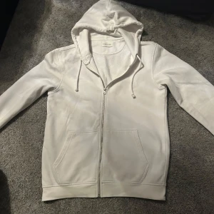 Vit hoodie från Pier One - Säljer en stilren vit hoodie från Pier One med dragkedja och justerbar huva. Perfekt för en avslappnad look. Den har långa ärmar och praktiska fickor framtill.