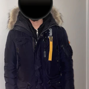 Svart parkajacka från Parajumpers - Säljer en marinblå/svart vinter parkajacka från Parajumpers med pälsfodrad huva och flera praktiska fickor. Jackan har en dragkedja och knappar, samt en gul remdetalj vid halsen också är avtagbar. Storlek M på jackan. Helt oskadad knappt använd. Vid intresse och mer, hör av dig 