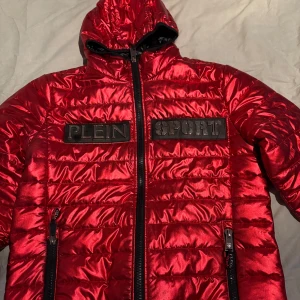 Plein jacka - Snygg röd quiltad jacka från Plein Sport med huva och dragkedja. Jackan har svarta detaljer och märkeslogga på bröstet. Perfekt för att hålla stilen under kyliga dagar.