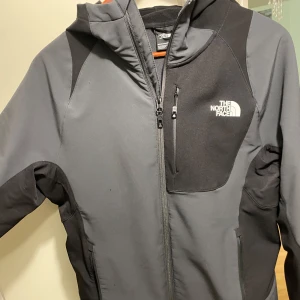 North Face höstjacka - Snygg grå och svart jacka från The North Face med huva och dragkedja. Perfekt för utomhusaktiviteter med praktiska fickor och en sportig design. Jackan har långa ärmar och är tillverkad i ett slitstarkt material.