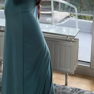 Blå axelbandslös långklänning - Elegant blå långklänning med en axelbandslös design och en smickrande slits. Klänningen har en draperad detalj vid midjan som ger en fin passform. Perfekt för speciella tillfällen.