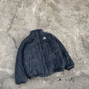 Svart pufferjacka från Adidas - Säljer en svart pufferjacka från Adidas med dragkedja och knappar. Jackan har  en hög krage för extra värme. Perfekt för kyliga dagar!!! Skulle säga att den passar mer M än L.  Mått: Längd 63 cm, bredd 52 cm