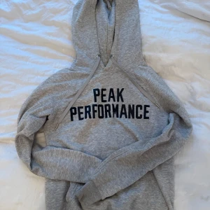 Grå hoodie från Peak Performance - Säljer en stilren grå hoodie från Peak Performance med svart text på bröstet. Den har en klassisk design med huva och dragsko.