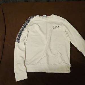 Vit tröja från Emporio Armani - Snygg vit tröja från Emporio Armani med EA7-logga på bröstet och längs ärmarna. Tröjan har en klassisk design med rund hals och långa ärmar. Perfekt för en stilren look. Kan sänka priset 