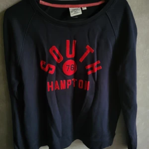 Mörkblå tröja från Crocker - Säljer en mörkblå tröja från Crocker med röd text 'South Hampton 76' på framsidan. Tröjan har långa ärmar och en rund halsringning. Perfekt för en avslappnad stil.