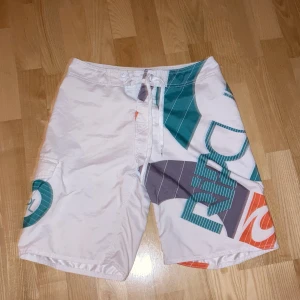 Rip curl shorts - Snygga vita badshorts från Rip Curl med färgglada grafiska mönster i turkos, orange och grått. Shortsen har en praktisk ficka på sidan och snörning i midjan för en justerbar passform. Perfekta för en dag på stranden eller vid poolen.