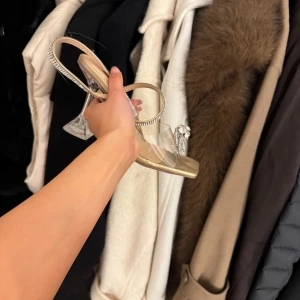 Eleganta klackskor med strassdetaljer - Snygga klackskor i guld med transparent rem och glittrande strassdetaljer. Perfekta för att ge en lyxig touch till din outfit. Klacken är genomskinlig och ger en modern känsla.