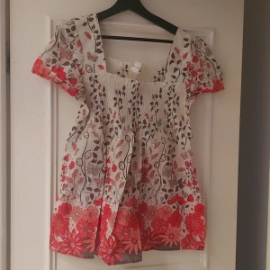Blommig topp med korta ärmar - Charmig topp med blommigt mönster i rött och beige. Den har korta ärmar och en smockad överdel som ger en bekväm passform. Perfekt för sommarens varma dagar.