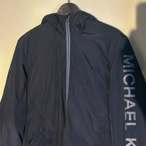 Mörkblå vindjacka från Michael Kors - Snygg mörkblå vindjacka från Michael Kors med dragkedja och huva. Logga längs ärmen och metall logga på höger axeln, perfekt för blåsiga dagar.