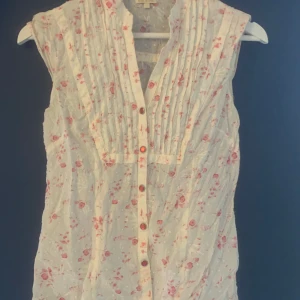 Sommrig blus - Elegant och bekväm blus från BikBok.  Denna ärmlösa blus har en vacker blommig design och röda knappar är perfekt för både vardagsbruk och festliga tillfällen. Tillverkad av mjuka material som ger en lätt och luftig känsla. Kombinera den med jeans eller en kjol för en stilren look. 