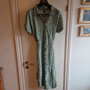 Grön blommig klänning från Shein - Säljer en söt grön klänning från Shein med ett blommigt mönster. Klänningen har korta ärmar och knappar framtill. Den har även ett knytband i midjan för en fin passform. Perfekt för en somrig look! 🌸