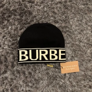 Burberry mössa - Snygg svart mössa från Burberry med vit text runt kanten. Perfekt för att hålla sig varm och stilren under kallare dagar. Märkets logga ger en exklusiv touch.
