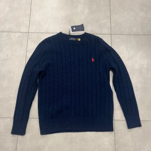 Mörkblå stickad tröja från Polo Ralph Lauren - Säljer en snygg mörkblå stickad tröja från Polo Ralph Lauren i storlek L med kabelstickat mönster och röd logga på bröstet. Tröjan har långa ärmar och ribbstickade muddar vid ärmslut och nederkant. Perfekt för kyliga dagar! 