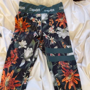 Blommiga leggings från Stronger - Snygga leggings från Stronger med ett färgglatt blommönster i orange, vitt och grönt. De har en hög midja med elastisk linning där 'STRONGER' är tryckt. Perfekta för träning eller en färgglad vardagslook.  Skriv via intresse och fler bilder!⭐️