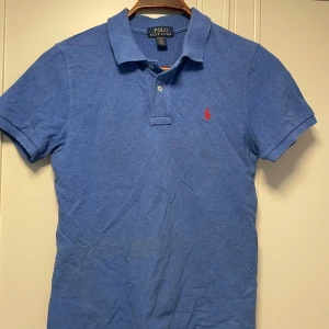 Blå pikétröja från Ralph Lauren - Säljer en klassisk mellanblå pikétröja från Ralph Lauren med röd logga på bröstet. Tröjan har korta ärmar och en stilren krage med knappar. Barnstorlek L = vuxenstorlek S. Prutat å klart!