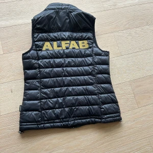 Svart quiltad väst med ALFAB-tryck - Säljer 3 st ALFAB västar 500 kr st (pris kan diskuteras) 2 st är i strl 36 och 1 är i strl 34. 2 st ät glansiga, och 1 är matt. Det står att den är såld men det är bara 1 av västarna som är sålda så det finns fortfarande 2 kvar!
