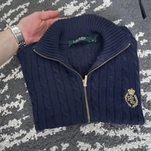Mörkblå stickad tröja från Ralph Lauren - Säljer en snygg mörkblå stickad tröja från Ralph Lauren med dragkedja och broderad logga på bröstet. Tröjan har en ribbad krage och långa ärmar, perfekt för kyligare dagar. Passar både till vardags och mer uppklädda tillfällen. Finns ett litet hål på framsidan (kolla bilder) inget märkbart men pga det lilla hålet är priset så lågt