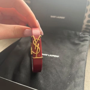 YSL armband  - Elegant rött armband från Saint Laurent med ett ikoniskt YSL-emblem i guld. Armbandet har en smal design och är tillverkat i läder med ett stilrent spänne. Tecken på användning, små slitage på spännet. Små färgningar på kanterna. Nypris 3000kr