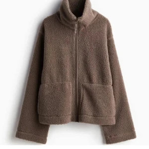 Brun teddyjacka med dragkedja - Mysig brun teddyjacka med hög krage och dragkedja framtill. Jackan har två stora fickor och är perfekt för kyliga dagar. Den har en avslappnad passform och är superbekväm att bära.