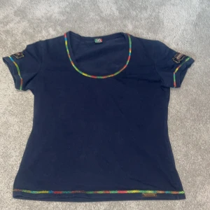 Mörkblå t-shirt med regnbågsdetaljer från Cappopera - Säljer en mörkblå t-shirt från Cappopera med färgglada regnbågsdetaljer längs halsringning, ärmslut och nederkant. T-shirten har korta ärmar och ett märke på ärmen. Perfekt för en färgglad touch i garderoben!