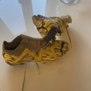 Fotbollsskor med kamouflagemönster - Snygga fotbollsskor med ett iögonfallande kamouflagemönster i gult och svart. Skorna har snörning och dobbar för bra grepp på planen. Perfekta för den som vill sticka ut på fotbollsplanen.