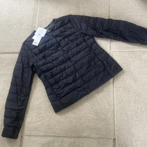 Svart dunjacka från Uniqlo - Säljer en stilren svart dunjacka från Uniqlo. Jackan är ultralätt och har en quiltad design med knappar framtill. Perfekt för kyliga dagar när du vill hålla värmen utan att känna dig tung. Ett måste i garderoben för den som gillar funktion och stil.