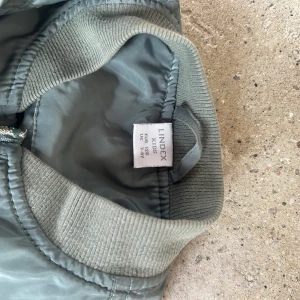 Grön bomberjacka från Lindex - Snygg grön bomberjacka från Lindex med dragkedja framtill och ribbade muddar vid ärmslut och nederkant. Jackan har praktiska fickor med knappar och en extra ficka med dragkedja på ärmen.