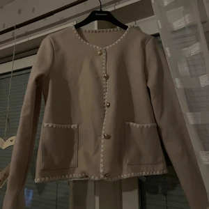 Beige kavaj med dekorativa knappar - Säljer en stilren beige kavaj med dekorativa knappar och vita detaljer längs kanterna. Kavajen har två framfickor och långa ärmar. Perfekt för en elegant look.