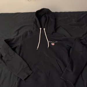 Svart hoodie från GANT - Säljer en stilren svart hoodie från GANT med broderad logga på bröstet. Den har en klassisk känguruficka och justerbar huva med vita snören. Perfekt för en avslappnad stil. Med storlek S. Pris kan diskuteras.