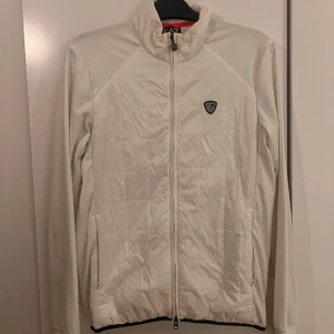 Vit jacka från Emporio Armani EA7 - Säljer en stilren vit jacka från Emporio Armani EA7. Jackan har en quiltad design med dragkedja framtill och ett EA7-märke på bröstet. Perfekt för en sportig look.