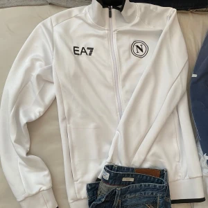 Vit jacka från EA7/napoli - Säljer en stilren vit jacka från EA7 med dragkedja och broderade detaljer. Jackan är fortfarande ny och har knappt använt den så de finns inga fläckar eller något sånt på den.