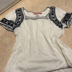 Vit blus med broderi från Indiska - Charmig vit blus från Indiska med svart broderi längs ärmar och hals. Blusen har korta ärmar och en luftig passform, perfekt för en bohemisk stil. Detaljerna ger en unik touch som gör den speciell.Den är helt ny, aldrig använd. Storlek L men sitter som M eventuellt lite oversize S. Fastpris 250kr