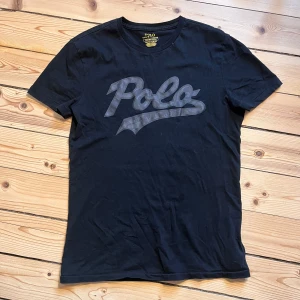Svart t-shirt från Polo Ralph Lauren - Säljer en svart t-shirt från Polo Ralph Lauren med ett stort Polo-tryck i grått på framsidan. T-shirten har en slim passform och är tillverkad i mjuk bomull. Perfekt för en stilren och avslappnad look. Sitter lite tight på mig som är 178 cm och 74 kg