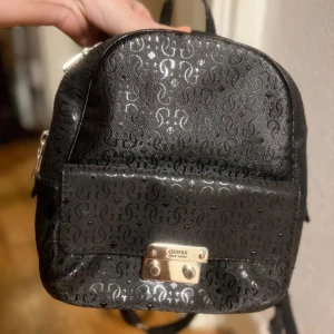 Svart ryggsäck från Guess - Snygg svart liten ryggsäck från Guess med ett subtilt mönster och guldfärgade detaljer. Väskan har en praktisk dragkedja och justerbara axelremmar. Perfekt för att bära dina dagliga nödvändigheter med stil.
