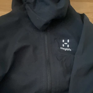 Svart vindjacka från Haglöfs - Säljer en svart vindjacka från Haglöfs med dragkedja framtill. Jackan har en enkel design med långärmade ärmar och en huva. Perfekt för blåsiga dagar och utomhusaktiviteter.