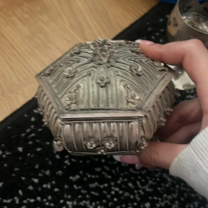 Antik smyckeskrin i silver - Vackert antikt smyckeskrin i silver med detaljerade ornament och en sexkantig form. Insidan är klädd i gul tyg, vilket ger en elegant kontrast till det silverfärgade yttre. Perfekt för att förvara små smycken eller andra värdefulla föremål.