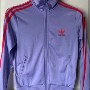 Lila jacka/tröja från Adidas - Säljer en snygg lila träningsjacka från Adidas med röda detaljer. Jackan har dragkedja framtill och två fickor med dragkedjor. Den ikoniska Adidas-loggan syns både fram och bak. Perfekt för en sportig look!