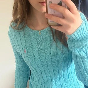Turkos stickad tröja från Ralph Lauren - Säljer en snygg turkos stickad tröja från Ralph Lauren med kabelstickat mönster. Tröjan har långa ärmar och en liten rosa logga på bröstet. Perfekt för en stilren look! 💙