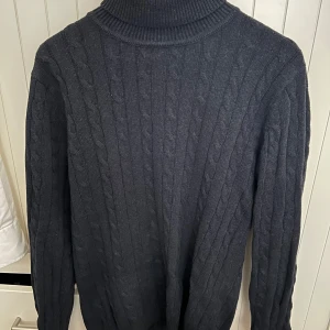 Mörkgrå stickad polotröja från John Henric - Säljer en stilren mörkgrå stickad polotröja från John Henric. Tröjan har ett klassiskt flätmönster och långa ärmar, perfekt för kyligare dagar. Den höga kragen ger extra värme och stil. Passar utmärkt till både jeans och chinos.