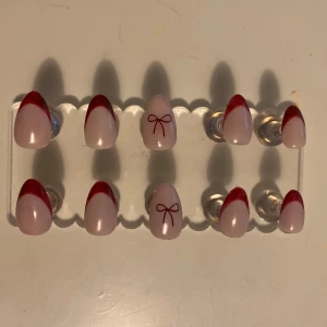 Red French tip (XXS-XS) Almond (lösnaglar💕) - Snygga press-on naglar med en elegant röd fransk manikyr och söt rosettdetalj på två naglar. Perfekt för att snabbt fixa en stilren look. Storlek är i XXS-XS!  Vid frågor eller intresse är du välkommen att kontakta mig!💕