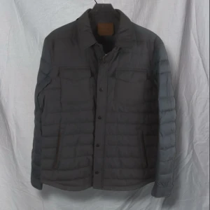 Grå quiltad jacka med svarta ärmar - Snygg grå quiltad jacka med svarta långärmade ärmar. Jackan har knappar framtill och två bröstfickor med lock. Perfekt för kyligare dagar. Från massimo dutti storlek L 