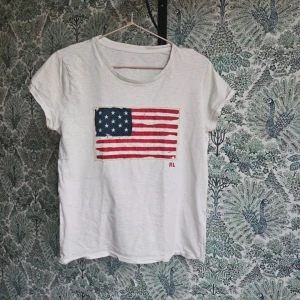 Vit ralph Lauren T-shirt  - Säljer en vit t-shirt från Ralph Lauren med en tryckt amerikansk flagga på framsidan. T-shirten har korta ärmar och en rund halsringning. Perfekt för en avslappnad stil.