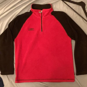 Umbro Quarterzip Fleece - Säljer en röd fleece tröja från Umbro med svarta ärmar och dragkedja. Perfekt för kyliga dagar. Tröjan har en hög krage och Umbro-loggan på bröstet. Använd men i fint skick!
