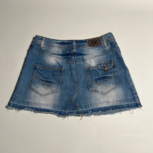 Lågmidjad jeanskjol  - Midja=37,5. Längd=33 cm. Snygg blå jeanskjol med slitningar och fransig kant. Kjolen har en klassisk femficksdesign och en knapp framtill. Perfekt för en avslappnad stil.