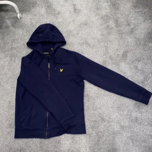 Mörkblå hoodie från Lyle & Scott - Snygg mörkblå hoodie från Lyle & Scott med dragkedja och huva. Den har en gul broderad logotyp på bröstet och är tillverkad i ett mjukt material. Perfekt för en avslappnad stil.