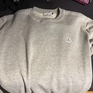 Grå Moncler tröja - Säljer en stilren grå sweatshirt från Moncler med rund hals och deras ikoniska logotyp på bröstet. Perfekt för en avslappnad och trendig look. Passar bra till både jeans och joggers.