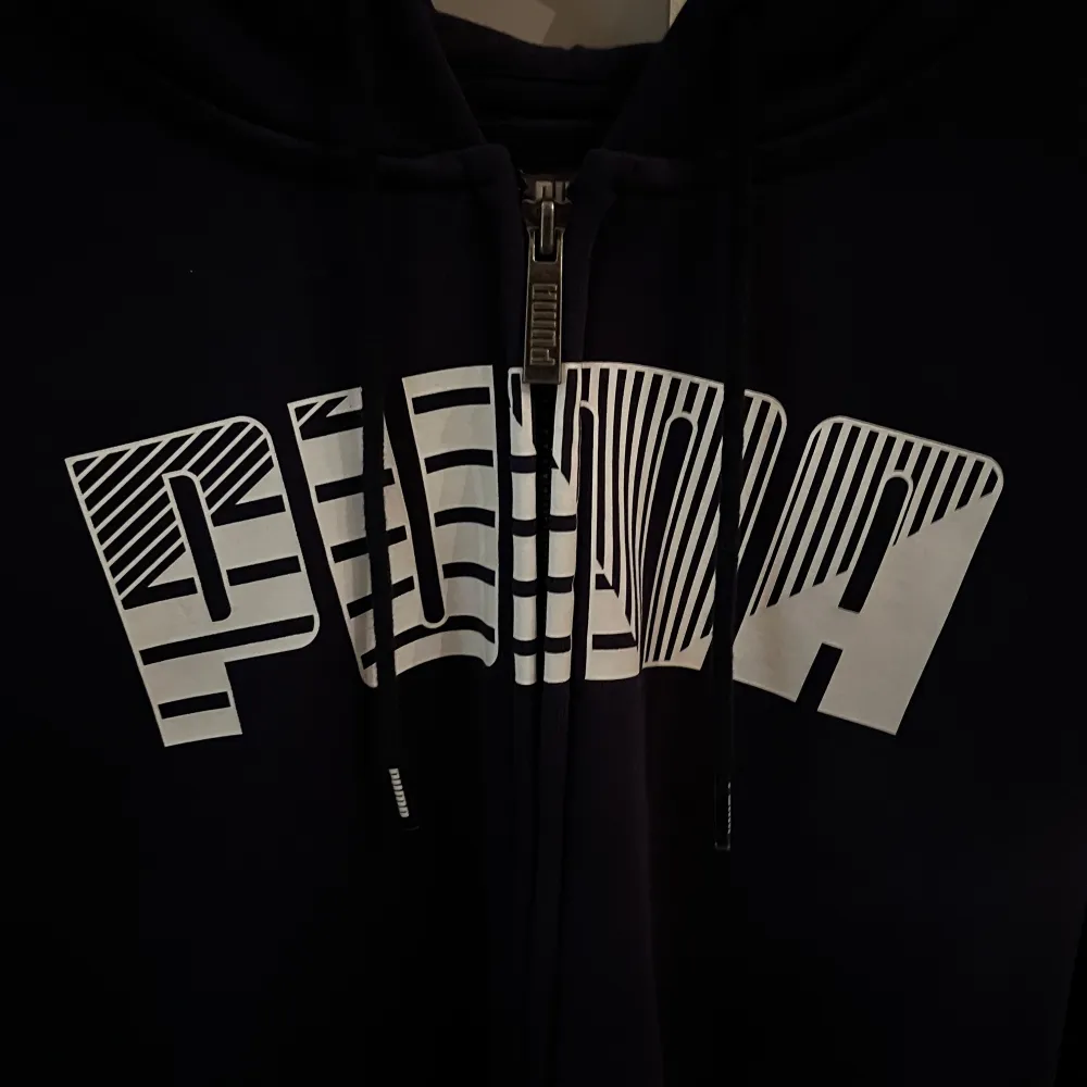 Säljer en svart hoodie från Puma med dragkedja och stor vit logga på framsidan. Perfekt för en avslappnad stil. Den har en bekväm huva med snörning och långa ärmar.. Neuletakit & Villapaidat.