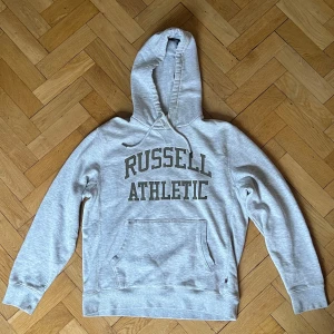 Grå hoodie från Russell Athletic - Säljer en klassisk grå hoodie från Russell Athletic med stor logga på framsidan. Tröjan har en bekväm passform med långa ärmar och en praktisk magficka. Perfekt för en avslappnad stil.